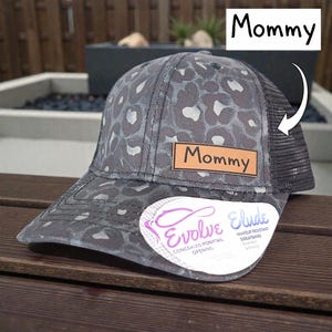 May include: Gray and black leopard print baseball cap with a mesh back. The cap features a brown leather patch with the word "Mommy" and a sticker that says "Evolve Elude".