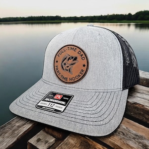 May include: A gray and black trucker hat with a brown leather patch that says "Full-Time Dad, Part-Time Hooker" with a fish illustration. The hat is adjustable and has a Richardson 112 label.