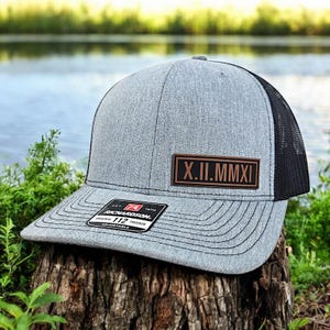 May include: A gray trucker hat with a black mesh back and a curved brim. A brown leather patch displays the Roman numerals X.II.MMXI. The hat has a Richardson 112 tag.