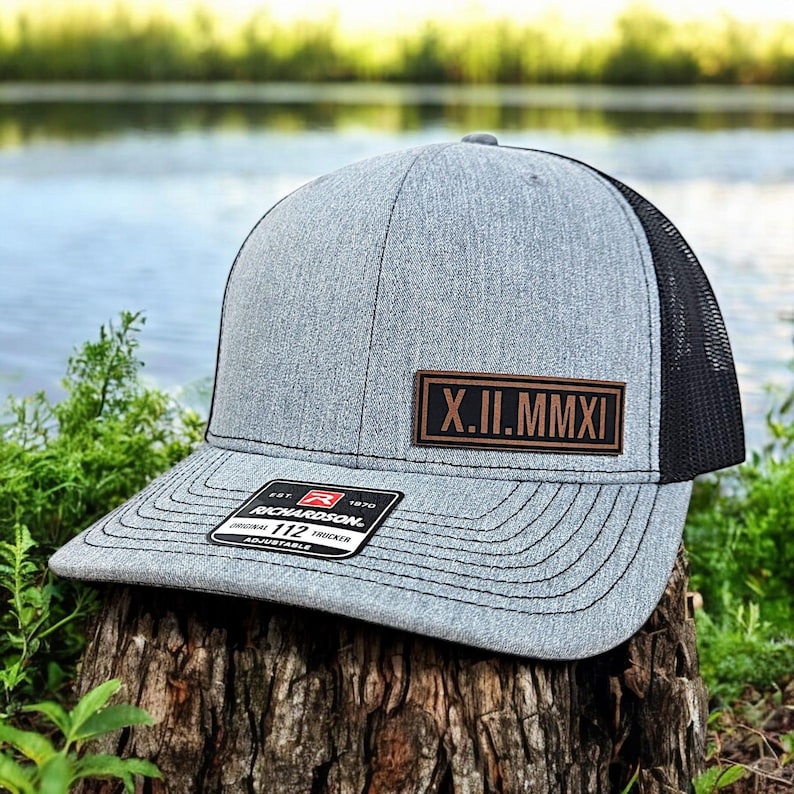 May include: A gray trucker hat with a black mesh back and a curved brim. A brown leather patch displays the Roman numerals X.II.MMXI. The hat has a Richardson 112 tag.