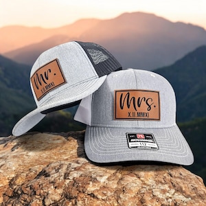 May include: Two gray trucker hats with leather patches. One hat reads "Mr. X.II.MMXI" and the other reads "Mrs. X.II.MMXI". The hats have a mesh back and a curved brim. The hats are on a rock with a mountain backdrop.