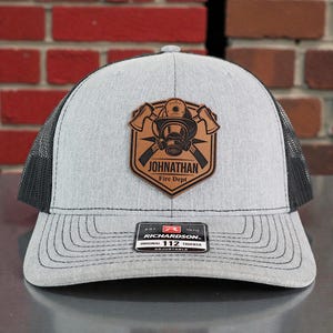 May include: A gray and black trucker hat with a leather patch featuring a firefighter's helmet, crossed axes, and the text "JOHNATHAN Fire Dept." The hat has a curved brim and an adjustable snap closure.