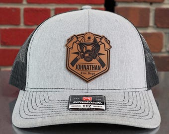 May include: A gray and black trucker hat with a leather patch featuring a firefighter's helmet, crossed axes, and the text "JOHNATHAN Fire Dept." The hat has a curved brim and an adjustable snap closure.