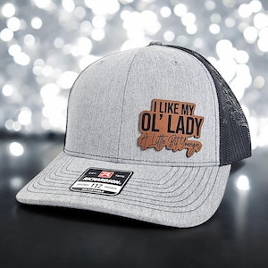 May include: A gray and black trucker hat with a brown leather patch that says "I like my ol' lady a little bit younger".