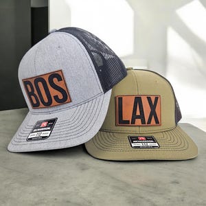 May include: Two trucker hats with black mesh back panels. The hat on the left is gray with a brown leather patch that says "BOS" in black letters. The hat on the right is olive green with a brown leather patch that says "LAX" in black letters.