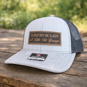 May include: A light gray trucker hat with a black mesh back and a curved brim. The hat features a brown rectangular patch with the text "I LIKE MY OL' LADY A Little Bit Younger". A small tag is visible on the brim.