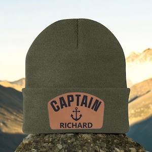 May include: Olive green beanie with a brown leather patch. The patch features the word "CAPTAIN" above an anchor symbol and the name "RICHARD" below. The beanie is made of a knitted material.