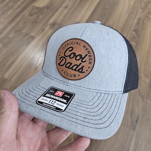 May include: Gray and black trucker hat with a brown leather patch that says "Official Member Cool Dads Club". The hat is adjustable and has a Richardson 112 label.