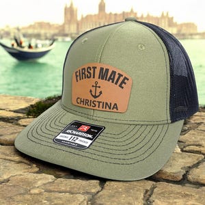 May include: Olive green trucker hat with a navy blue mesh back. The hat features a brown leather patch with the words "FIRST MATE" and "CHRISTINA" along with an anchor graphic. The hat is adjustable and has a curved brim.
