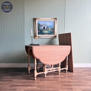 May include: A light brown, antique drop-leaf table with a rounded edge. The table has four legs and a supporting structure. A painting in a gold frame and a candle holder are on the wall behind the table. The wall is painted a light green color.
