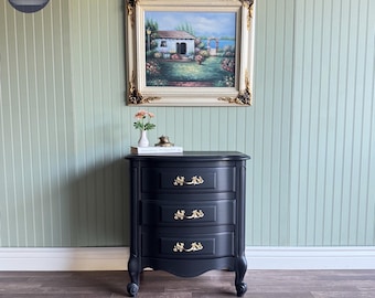 May include: Black three-drawer nightstand with gold-colored hardware. The nightstand has a curved front and sits on four curved legs. A framed painting hangs above the nightstand. The painting depicts a house and garden scene.