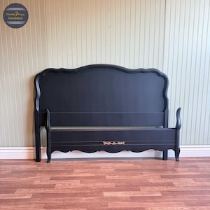 May include: Black wooden bed frame with a decorative headboard and footboard. The headboard has a curved top edge. The footboard has a decorative gold accent. The bed frame is on a wood floor.