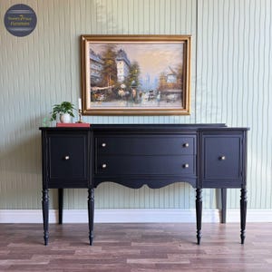 May include: A black buffet with two drawers and two cabinet doors, featuring gold-colored knobs. The buffet has ornate legs and is topped with a framed painting of a street scene. A small plant and candle holder are on top.