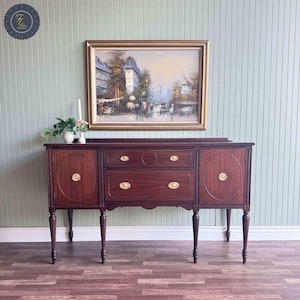 May include: A dark mahogany wood sideboard with brass hardware and four legs. The sideboard has two doors on the left, two drawers in the center, and two doors on the right. The sideboard is in front of a light green wall with white trim.