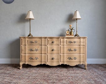 May include: A light brown wooden dresser with nine drawers featuring ornate metal pulls. Two brass lamps with off-white shades sit on top, alongside a small clock and a decorative pitcher. The text Twenty 7th Ave. Furniture is visible.