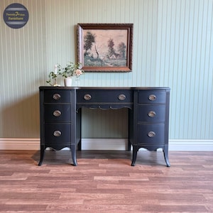 May include: A black vintage desk with three drawers on each side and a center opening. The desk has brass-colored hardware and a scalloped edge. A framed landscape painting hangs above the desk.