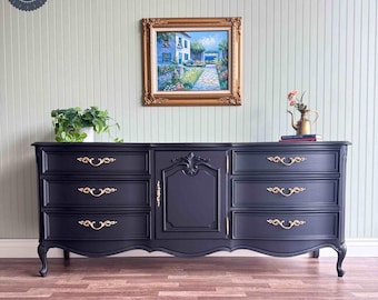 May include: A black painted dresser with six drawers and two doors. The dresser has gold-colored hardware and sits on a dark wood floor. A painting of a house with a garden is hanging on the wall behind the dresser.