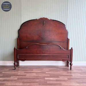 May include: A dark cherry-stained wooden bed frame with a high, ornate headboard. The headboard features a curved top with decorative carvings and a lower, arched section. The footboard mirrors the design. The bed frame is a classic piece of bedroom furniture.