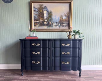 May include: A black dresser with six drawers and gold handles. The dresser has a curved front and sits on short legs. The dresser is in front of a green wall with a painting of a city scene above it.