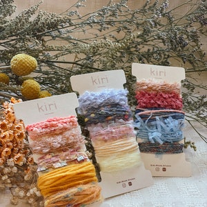 Pode incluir: Três rolos de fio colorido com o texto "kiri made studio" em cada etiqueta. O fio é uma mistura de texturas, incluindo felpudo, felpudo e brilhante.