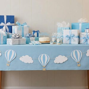 May include: A light blue tablecloth decorated with a garland of white clouds and hot air balloons. On the table are various gift boxes wrapped in blue, white, and silver paper with ribbons, alongside a small round cake and treat bags.