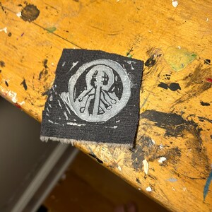 May include: A black fabric patch with a white circular design featuring a stylized tree with branches and leaves. The design is centered on the patch.