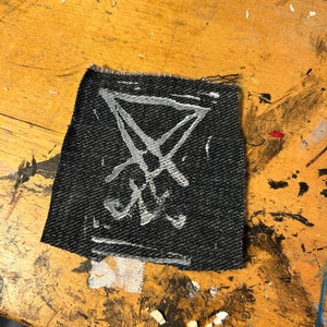 Puede incluir: Un parche de tela negro con un s&iacute;mbolo de pentagrama blanco dibujado en &eacute;l.