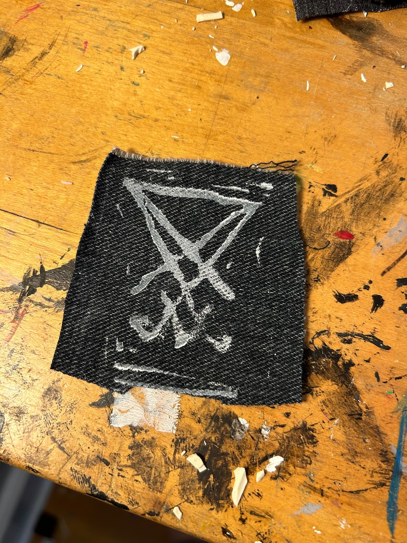 Puede incluir: Un parche de tela negro con un s&iacute;mbolo de pentagrama blanco dibujado en &eacute;l.