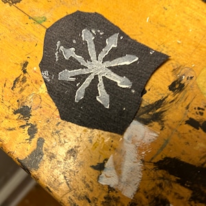 May include: A black fabric patch with a white eight-pointed star design. The star has a jagged, hand-drawn look.