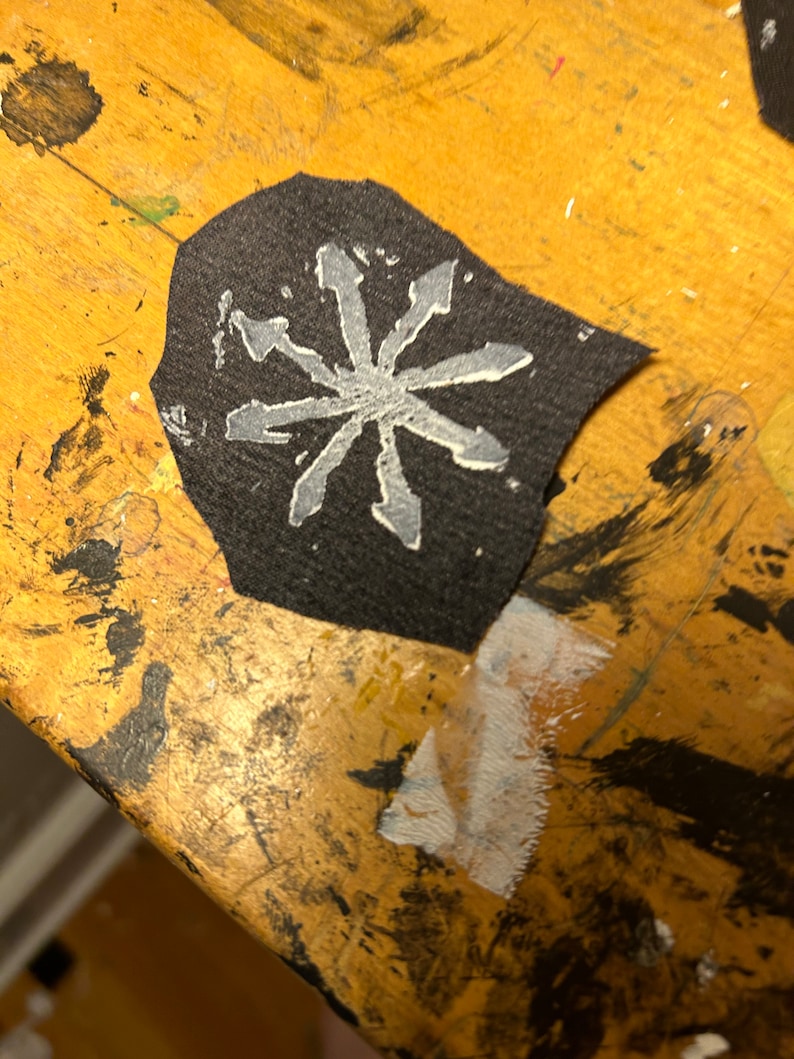 Puede incluir: Un parche de tela negro con un dise&ntilde;o de estrella blanca de ocho puntas. La estrella tiene un aspecto irregular, dibujado a mano.