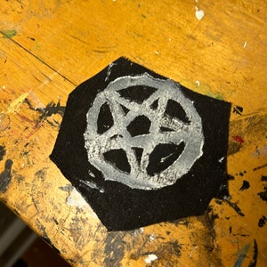 Puede incluir: Un parche de tela negro con un dise&ntilde;o de pentagrama blanco. El parche est&aacute; sobre una superficie de madera.