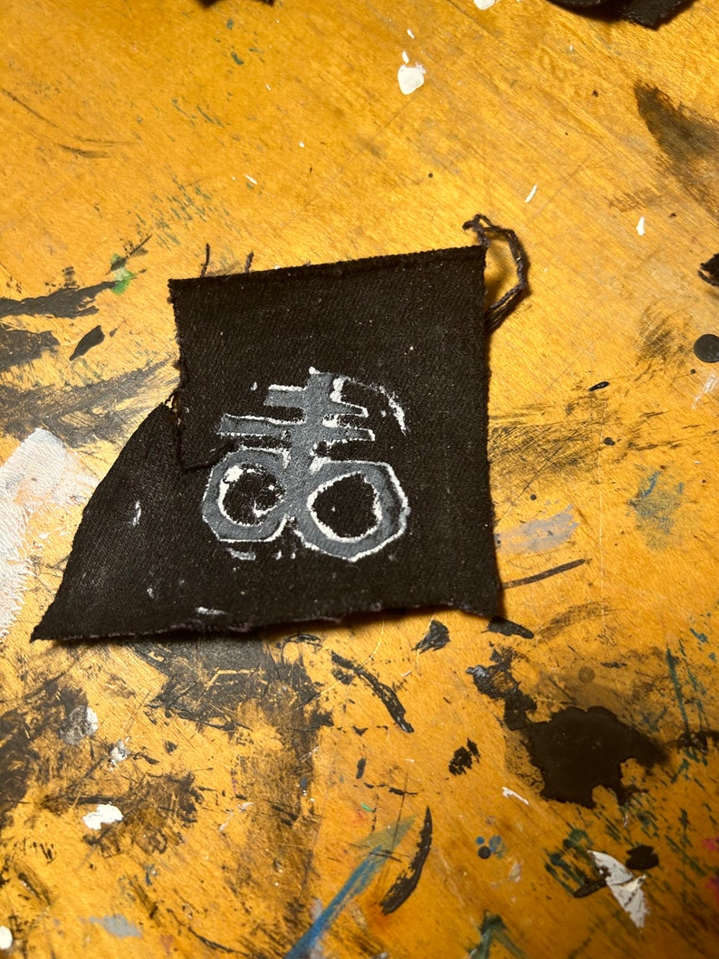 Puede incluir: Un parche de tela negro con un car&aacute;cter kanji blanco en &eacute;l. El car&aacute;cter es una versi&oacute;n estilizada de la palabra japonesa "夢" (yume), que significa "sue&ntilde;o".