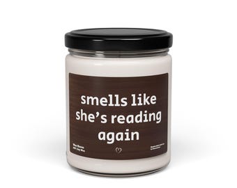 May include: A white candle in a glass jar with a black lid. The candle has a brown label with the text "smells like she's reading again" in white. A small heart is below the text.