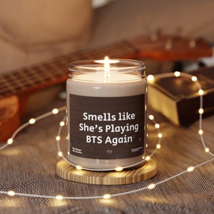 May include: A brown candle with a white label that reads "Smells like She's Playing BTS Again". The candle is lit and sitting on a wooden coaster.