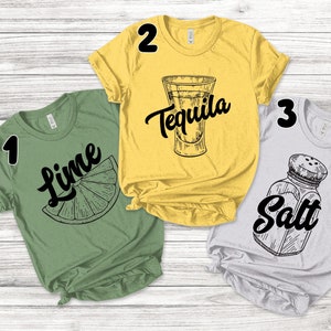 May include: Three short-sleeved t-shirts with black graphics on a light wood background. The shirts are green, yellow, and grey. The green shirt has a lime slice and the word "Lime". The yellow shirt has a tequila shot and the word "Tequila". The grey shirt has a salt shaker and the word "Salt".