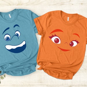 May include: Two t-shirts with cartoon faces printed on them. The blue shirt has a blue face with a surprised expression and the orange shirt has an orange face with a smiling expression.