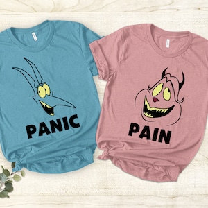 May include: Two t-shirts, one light blue and one pink, with cartoon characters and text. The blue shirt has a blue and yellow cartoon character with the text "PANIC". The pink shirt has a pink and yellow cartoon character with the text "PAIN".