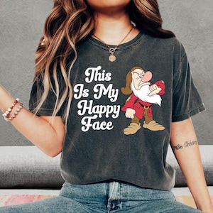 以下が含まれることがあります： 暗いグレーのTシャツに、赤い服を着た不機嫌そうなドワーフの白いグラフィックがプリントされています。Tシャツには「This Is My Happy Face」という白い文字がプリントされています。