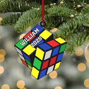 Puede incluir: Adorno navideño de Rubik's Cube con el nombre "WILLIAM" y el año "2025" impresos. El adorno cuelga de una cinta roja sobre un fondo de árbol de Navidad.