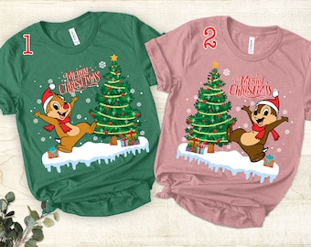 May include: Two short-sleeved t-shirts, one green and one pink, feature a cartoon character in a Santa hat and scarf, sliding on snow next to a Christmas tree. The text "Merry Christmas" is above the tree.