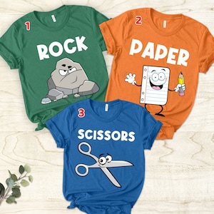 Puede incluir: Tres camisetas con un tema de piedra, papel o tijeras. La camiseta verde tiene una piedra de dibujos animados con una cara y la palabra "ROCK". La camiseta naranja tiene un papel de dibujos animados con una cara y la palabra "PAPER". La camiseta azul tiene unas tijeras de dibujos animados con una cara y la palabra "SCISSORS".