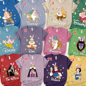 May include: A set of 12 t-shirts featuring different characters from the Disney movie Snow White and the Seven Dwarfs. Each shirt has a different character with a bunny ear design and the character's name printed on the shirt. The shirts are in various colors, including blue, purple, pink, red, green, and beige.