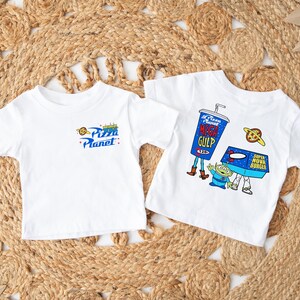 May include: Two white t-shirts with short sleeves. One displays the "Pizza Planet" logo in blue, yellow, and red. The other features a "Mega Gulp" cup, a "Super Nova Burger" box, and an alien character.