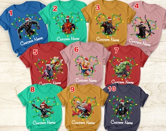 May include: Ten different t-shirts with various superhero characters wearing Santa hats and surrounded by Christmas lights. The shirts are all different colors and have the text "Custom Name" printed on them.