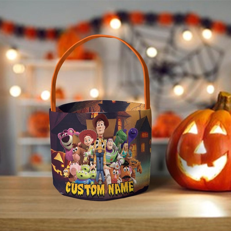 May include: A cylindrical Halloween trick-or-treat bucket with an orange handle, featuring a colorful graphic of Toy Story characters including Woody, Buzz Lightyear, and Jessie against a spooky house background with the text CUSTOM NAME.