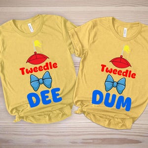 Könnte beinhalten: Zwei gelbe T-Shirts mit den Wörtern "Tweedle Dee" und "Tweedle Dum" in Rot und Blau, mit einem roten Regenschirm und einer blauen Fliege. Die Shirts sind aus weichem Material.