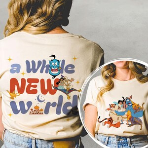 May include: Cream-coloured t-shirt with a colourful graphic from the film Aladdin. The design includes the text "a whole new world" and illustrations of characters like Genie, Aladdin, and Jasmine. The shirt has a crew neck and short sleeves.