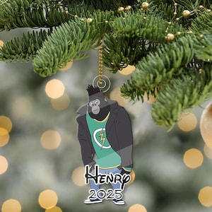 May include: A clear acrylic Christmas tree ornament depicting a cartoon gorilla in a black jacket and green shirt with a lightning bolt. The name "Henry" and the year "2025" are printed below. The ornament hangs from a gold chain.