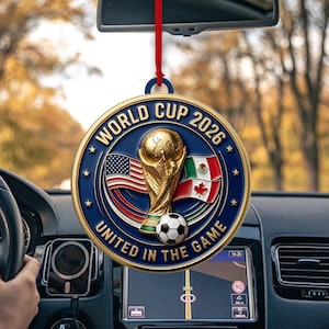 May include: A round, gold-rimmed hanging ornament featuring a blue center with the text WORLD CUP 2026 UNITED IN THE GAME. The design includes a gold trophy, a soccer ball, and the flags of the United States, Mexico, and Canada, suspended by a red cord.