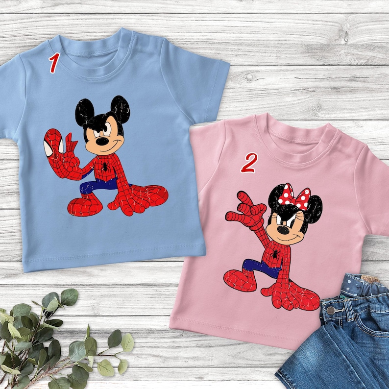 May include: A light blue t-shirt featuring a graphic of Mickey Mouse in a red and blue spider-themed costume and a pink t-shirt featuring a graphic of Minnie Mouse in a red and blue spider-themed costume, both labeled with the numbers 1 and 2.